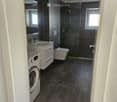 Modern bathroom with dark grey tiles, a white sink, toilet, shower, and a private washing machine.