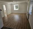 Empty room with wooden flooring, white walls, and a window with a view of greenery. The room features a radiator on one wall and a door leading to another space.