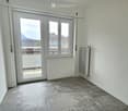 Empty room with grey tiled floor, white walls, large sliding glass door leading to a balcony, and radiators on both sides of the door.