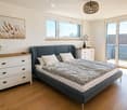 Modern bedroom with a grey upholstered bed, white pillows, a wooden nightstand, a drawer unit, potted plants, and a large window with a view to a balcony.