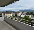 Balcony with white railing, overlooking a residential area with houses, trees, and distant mountains.