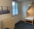 A small bedroom with a single bed, a wooden cabinet, a radiator, and three framed mirrors on the wall. The room has a window with white curtains and a pendant light fixture.