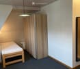 Small bedroom with a single bed, a small stool, and a wooden wardrobe. The room has a slanted ceiling and is well-lit with a hanging light fixture.