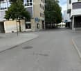 Urban street scene with modern buildings, trees, and parking signs. The road is empty, and there are no visible vehicles or pedestrians.