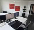 Modern office space with a white desk, ergonomic chair, dual monitors, laptop, keyboard, mouse, and a potted plant. The room features white walls, black flooring, and abstract art pieces.