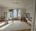 Bright, airy room with wooden herringbone flooring, large white rug, large windows with open shutters, and various children's furniture and toys.