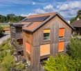 Modern two-story house with wooden exterior, solar panels on the roof, and a balcony with outdoor furniture.