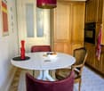 A cozy kitchen with a round white dining table, red chairs, a wicker chair, a bowl of fruit, a pepper grinder, wooden cabinets, and an oven.