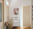A narrow hallway with wooden flooring, a hanging spherical light fixture, a small white cabinet with glass top, a round rug with floral pattern, and a folding chair.