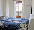 Cozy bedroom with a large bed covered in a floral-patterned duvet, a nightstand with a lamp, a wooden door leading to a balcony, and a small armchair with a patterned pillow.