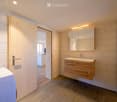 Modern bathroom with light wooden cabinet, sink, and mirror. The room features light-colored tiles and a wooden door leading to another space.