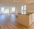 Empty kitchen with light wooden flooring, white walls, and a modern kitchen island with a sink and faucet. Large windows provide natural light.