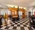 A spacious, elegantly furnished room with a black and white checkered marble floor, multiple tables and chairs, a refrigerator, wooden cabinets, and large windows with curtains.