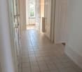 A clean, tiled hallway with white walls, multiple doors, and a slanted wooden ceiling. Natural light streams in from an open doorway at the end.