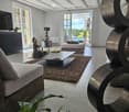 Modern living room with large windows and a view, featuring a sofa, coffee table, ottoman, floor lamp, and decorative sculpture.