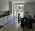 Open-plan kitchen and dining area with white cabinets, a dark dining table with chairs, and a vase of flowers on the table. The kitchen features a sink, oven, and dishwasher, with a view to a garden through large glass doors.