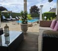 Outdoor patio with wooden decking, glass table, wicker sofa, lantern, potted plant, and view of swimming pool.