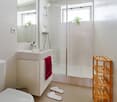Modern bathroom with white tiles, a sink, mirror, toilet, glass-enclosed shower, a red towel, a wooden shelf, and two white bathroom slippers on the floor.