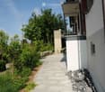A well-maintained garden area with a paved pathway leading to a modern building. The garden features neatly trimmed bushes, small trees, and a stone wall. A balcony with a metal railing is visible on the building.