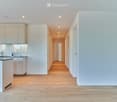 Bright, modern hallway with light wooden flooring, white walls, and recessed lighting. The hallway leads to a kitchen area with white cabinets, a countertop, and a sink.