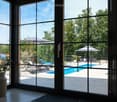Bright room with large glass doors opening to a patio with a swimming pool, lounge chairs, and umbrellas. A vase with flowers is placed on a table near the glass doors.