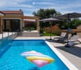 A modern outdoor area features a swimming pool with inflatable toys floating in it, lounge chairs under an umbrella, a patio with a dining set, and a house with a covered walkway.