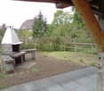 Outdoor patio area with a stone barbecue, seating benches, and a wooden fence, surrounded by greenery and a house in the background.