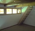 An attic space with exposed wooden beams, a wooden staircase leading up, and several windows letting in natural light. The room is empty with no furniture or furnishings.