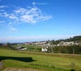 A panoramic view of a rural village nestled in a green valley, with scattered houses, trees, and open fields under a clear blue sky.