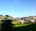 A panoramic view of a town with a mix of residential and commercial buildings, surrounded by green hills and a clear blue sky. A prominent aqueduct structure is visible in the distance.
