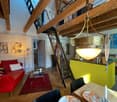 Open-plan living and dining area with exposed wooden beams, a red sofa, a glass coffee table, a dining table set for two, and a bright yellow kitchen area.
