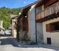 A row of traditional wooden and stone houses in a narrow village street, surrounded by greenery and mountains.