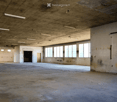 Empty industrial-style room with concrete floors and walls, large windows, and exposed ceiling with fluorescent lighting.