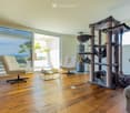 Bright living room with large windows offering a scenic ocean view, featuring a cream-colored sofa, two white armchairs, a multi-level cat tree, a small coffee table with a chessboard, and a polished wooden floor.