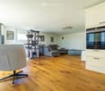Bright, open living space with light-colored walls and wooden flooring. Features a grey sofa, a single chair, a cat tree, a coffee table, and a modern oven with cabinets above.