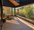 Wooden balcony with a modern design, featuring a lounge chair, small table, potted plants, and a scenic countryside view.