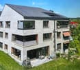 Modern two-story house with grey concrete facade, multiple windows, balconies, and a well-maintained garden area with outdoor furniture.