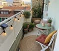 Cozy balcony with a round jute rug, white folding chair with red cushion, small green table, potted plants, string lights, and a view of the city.