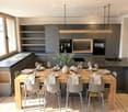 Modern kitchen with grey cabinets, wooden dining table, and grey chairs. Features a wine cooler, sink, and decorative vase with greenery.