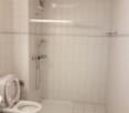 Small bathroom with white tiled walls and floor, featuring a toilet, a shower area with a handheld showerhead, and a small shelf above the shower.