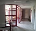 A well-lit hallway with tiled flooring, white walls, and red-framed glass doors leading to a room with wooden chairs and a table. A fire extinguisher is mounted on the wall.