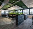 Modern office space with desks, chairs, computers, and large windows offering a scenic view. Features include grey carpeting, grey cabinets, and indoor plants.