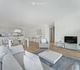 Modern, open-concept living and dining area with light grey furniture, white coffee table, grey sofa, armchair, dining table with chairs, and a television on a stand. The space features light wood flooring, white walls, and contemporary decor including po