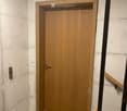 A narrow hallway with a wooden door and a staircase railing visible. The walls are concrete, and the floor is tiled.
