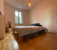 Cozy bedroom with wooden flooring, large window, bed with pillows and blankets, nightstand, and decorative vase.