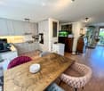 Open-plan kitchen and living area with modern white cabinets, a wooden dining table with chairs, a fish tank, and a cat tree. The kitchen features stainless steel appliances, including a refrigerator, oven, and microwave, and a countertop with various ite