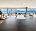 Modern open-plan living and dining area with large windows offering a scenic mountain view, featuring a white dining table, grey chairs, a white sofa, a wooden coffee table, and several potted plants.