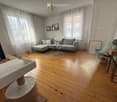 Modern living room with wooden flooring, large windows, grey sectional sofa, coffee table, cat tree, and decorative items.