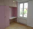 Empty room with light pink walls, wooden floor, built-in cabinets, and a window with a view of the street.