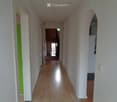 A long, narrow hallway with light wooden flooring and white walls. The hallway features several doors on both sides, with one door painted green. An arched opening leads to another room at the end of the hallway.